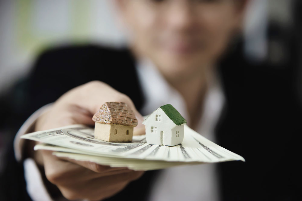 Una ejecutiva extendiendo si mano con unos billetes y sobre ellos unas mini casas de juguete